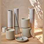 A collection of Skeem Design citronella products on a wooden table featuring matte beige ceramic containers with embossed insect motifs like mosquitoes, dragonflies, and moths, cylindrical tubes with colorful Mojave Suede and Cypress Drift labels, and an incense stick in a small dish under dappled leaf shadows