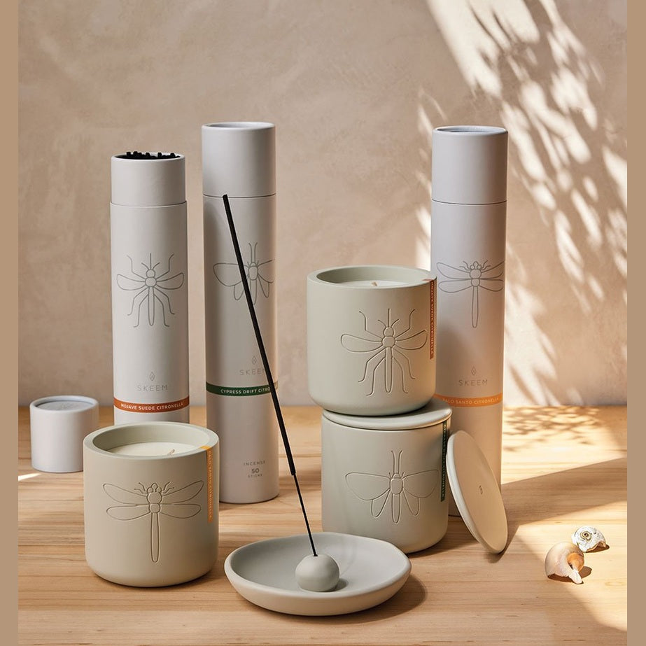 A collection of Skeem Design citronella products on a wooden table featuring matte beige ceramic containers with embossed insect motifs like mosquitoes, dragonflies, and moths, cylindrical tubes with colorful Mojave Suede and Cypress Drift labels, and an incense stick in a small dish under dappled leaf shadows