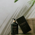 L'AVANT Collective matte black Fresh Linen hand soap bottle with a gold pump and textured base stands beside its matching black box with gold lettering, both on a light wooden surface with a palm leaf shadow cast on the wall