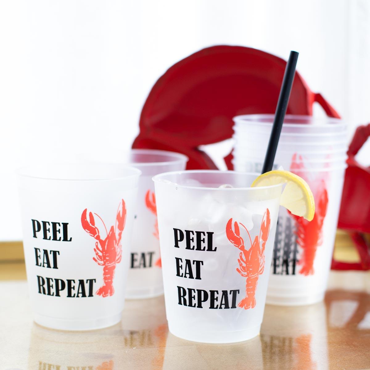 Frosted disposable party cups adorned with a vibrant red lobster graphic and "PEEL EAT REPEAT" text, one cup holding ice with a fresh lemon slice and black straw, set with matching red lobster plates in the background