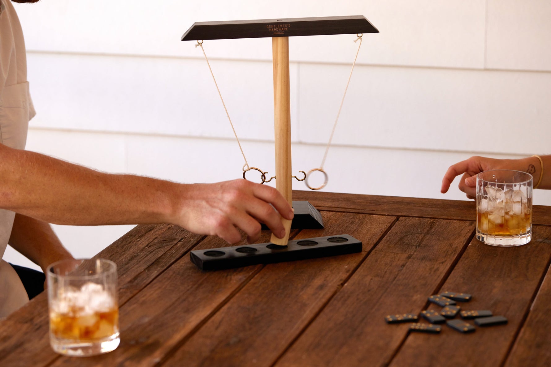 Photograph depicting a hand playing a Gentlemen's Hardware ring toss game on a rustic wooden outdoor table, beside two whiskey glasses with ice and scattered dominoes with gold dots