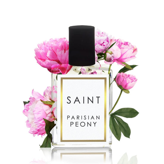A clear glass SAINT Parisian Peony perfume bottle with a black cap and a white label with gold trim, elegantly surrounded by vibrant pink peonies and lush green leaves