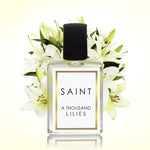 Digital product photograph of a Saint A Thousand Lilies clear glass perfume bottle with a black cap and gold-bordered white label centered against a backdrop of fresh white lilies with striking yellow-orange stamens casting a subtle reflection below