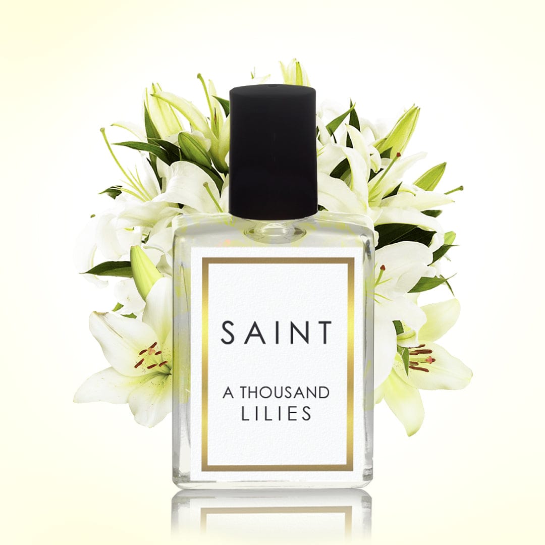 Digital product photograph of a Saint A Thousand Lilies clear glass perfume bottle with a black cap and gold-bordered white label centered against a backdrop of fresh white lilies with striking yellow-orange stamens casting a subtle reflection below
