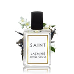 Product photograph of a clear rectangular perfume bottle with a black cap labeled SAINT JASMINE AND OUD with a gold border surrounded by delicate white jasmine flowers and dark oud wood chips on a reflective surface