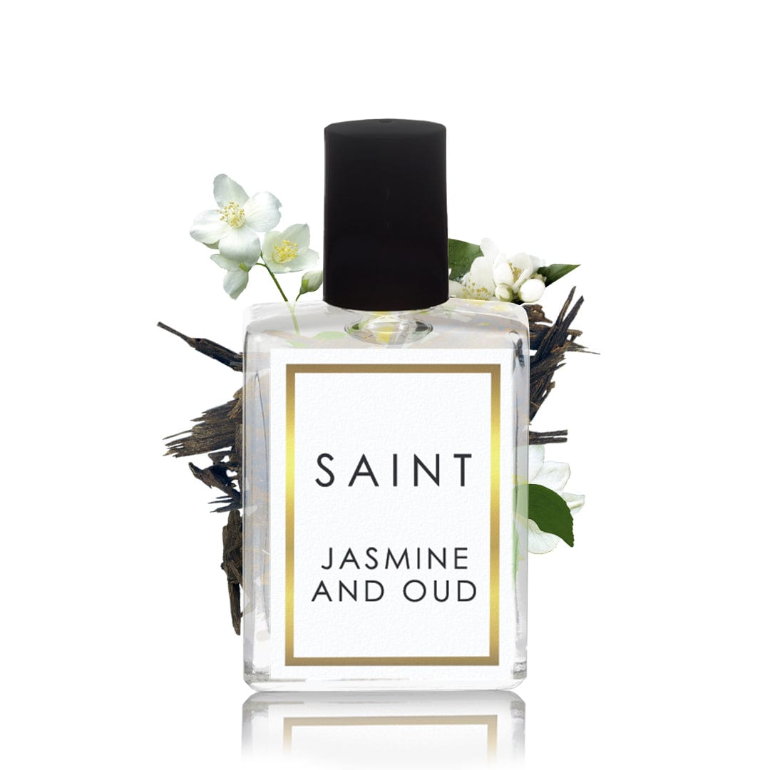 Product photograph of a clear rectangular perfume bottle with a black cap labeled SAINT JASMINE AND OUD with a gold border surrounded by delicate white jasmine flowers and dark oud wood chips on a reflective surface