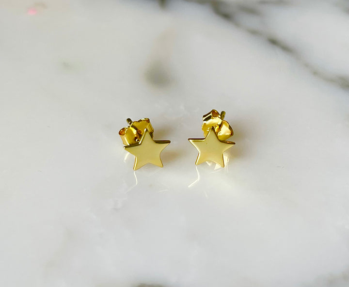 A pair of shiny gold star stud earrings with a minimalist design resting on a smooth white marble surface