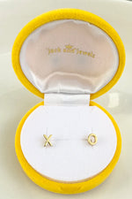 Product photography featuring Jack and Jewelz gold X and O stud earrings nestled in a bright yellow velvet jewelry box with a pristine white satin lining