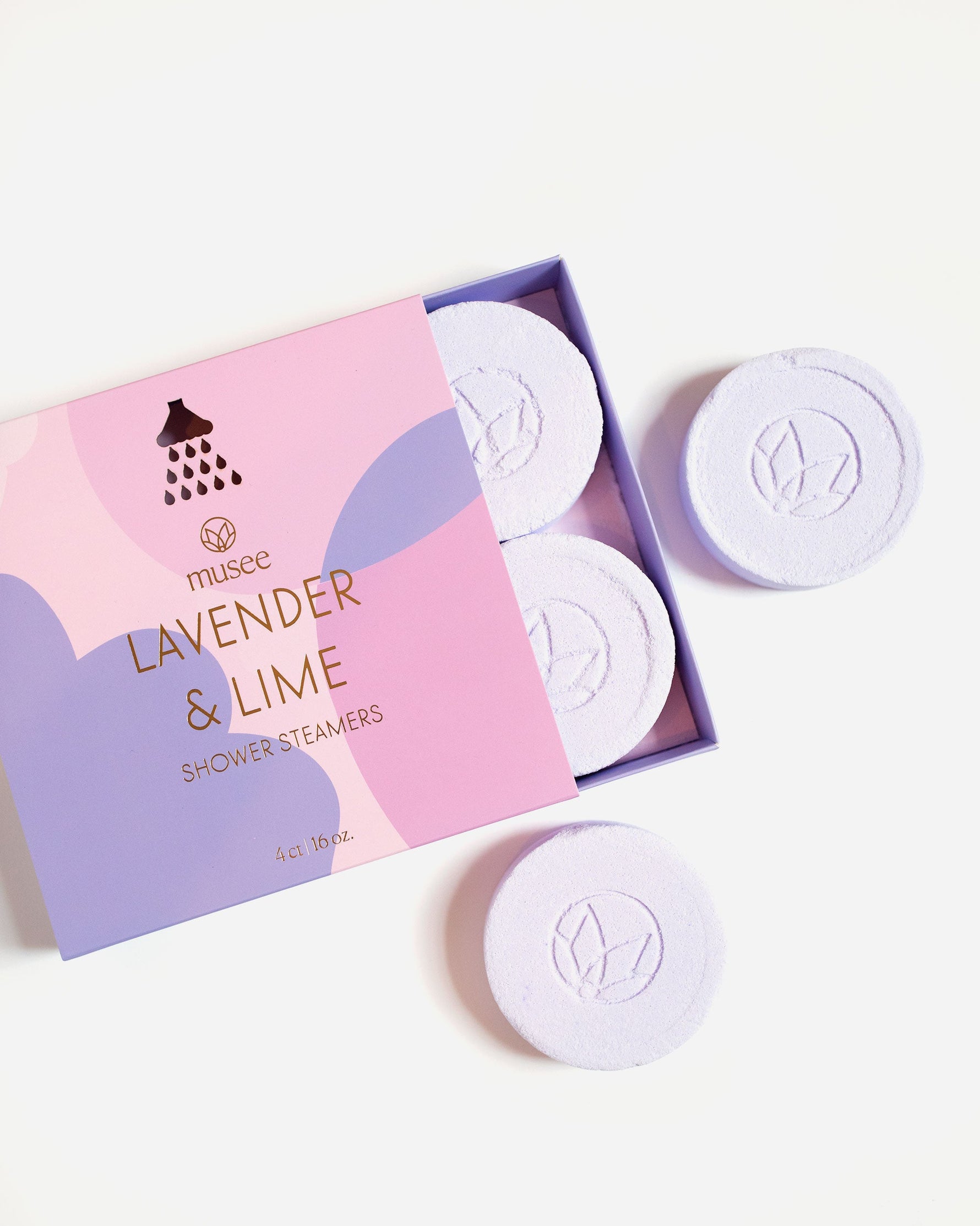 Product photography of Musee Lavender & Lime Shower Steamers, featuring a pink and purple box with gold text and showerhead icon, revealing light purple embossed circular steamers on a white background
