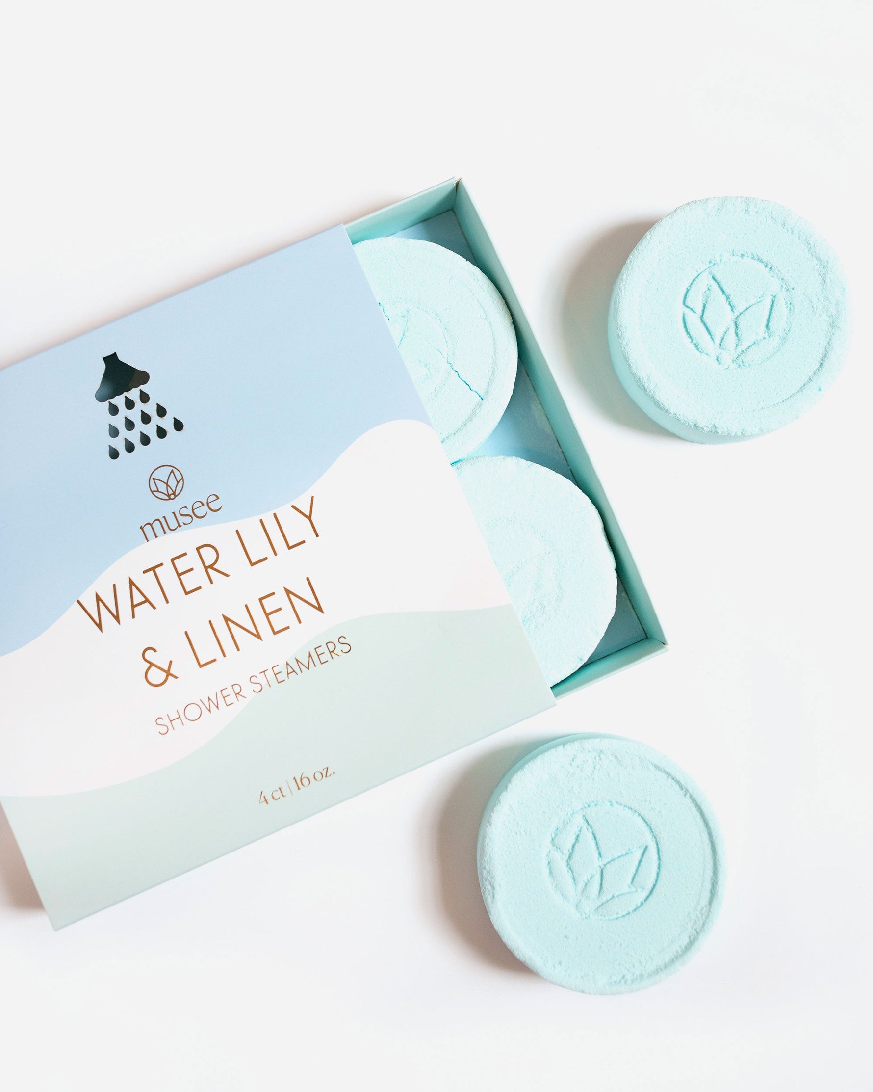 Musée Water Lily & Linen Shower Steamers in a pastel blue box featuring a showerhead icon and brown lettering, displaying four round light blue fizzers embossed with the brand's leaf logo, two inside and two on a clean white background