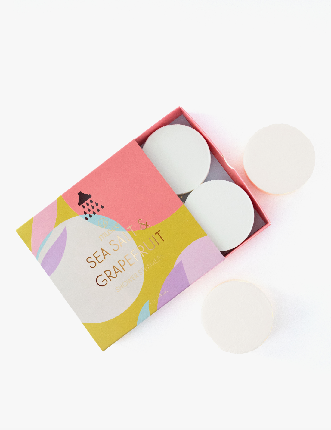An open pink and yellow box of Sea Salt & Grapefruit shower steamers featuring a black shower graphic and abstract pastel shapes rests on a white surface with three white circular steamers, two inside and one beside it
