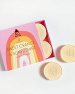 Musee product image featuring Sweet Orange & Sunflower Shower Steamers in a vibrant pink box with a rainbow design and red showerhead icon, showcasing gold foil text and pale yellow round steamers embossed with the Musee logo on a clean white background
