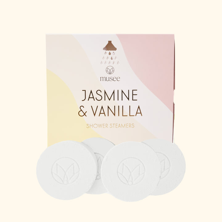 Product photograph by Musee showing Jasmine & Vanilla Shower Steamers in a pastel pink and yellow wavy box featuring a showerhead icon, alongside four white circular steamers with an embossed leaf logo