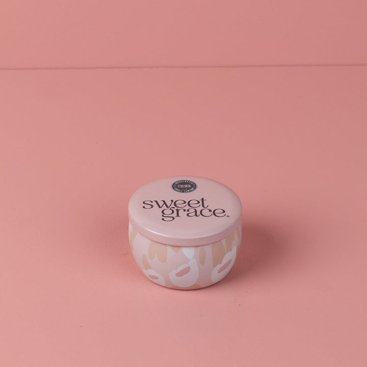 Pale pink Sweet Grace candle tin featuring white and tan abstract patterns on its body and black text on its lid, set against a vibrant pink background