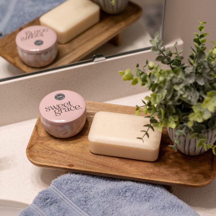 A close-up of bath essentials on a white speckled bathroom counter, displaying a pink "Sweet Grace" candle and a creamy bar of soap on a wooden tray, with a vibrant green artificial plant and a soft blue towel