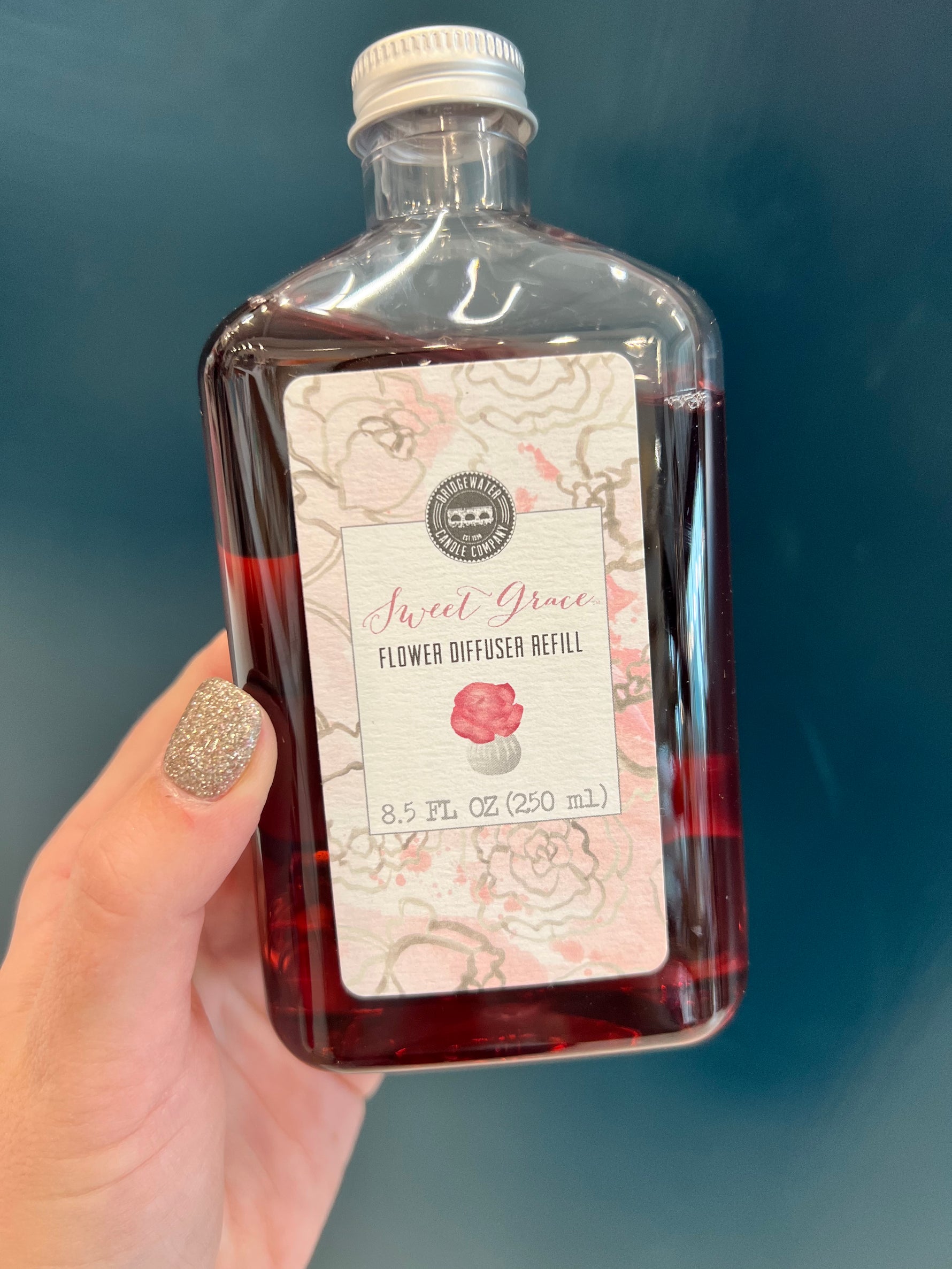 A bottle of Sweet Grace Flower Diffuser Oil Refill with a label in front, on a blue background