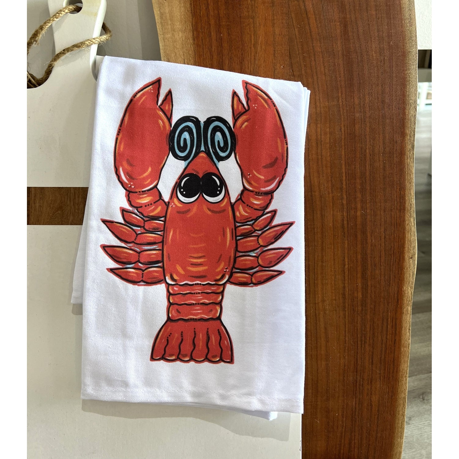A white cotton tea towel with a cartoon illustration of a red crawfish in the center, displayed on a wooden surface.