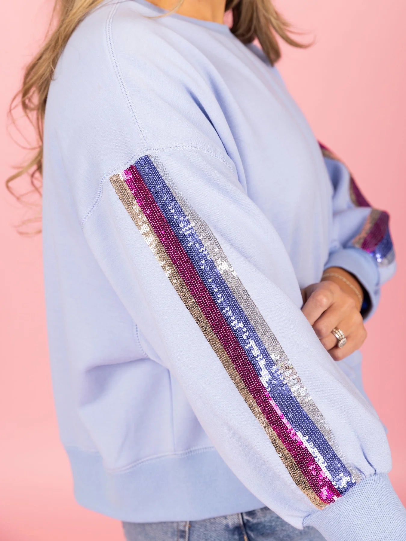 Light blue crewneck sweatshirt featuring multi-colored sequin stripes in shades of gold, silver, pink, and purple on the sleeves worn by a woman with a ring against a vibrant pink studio background