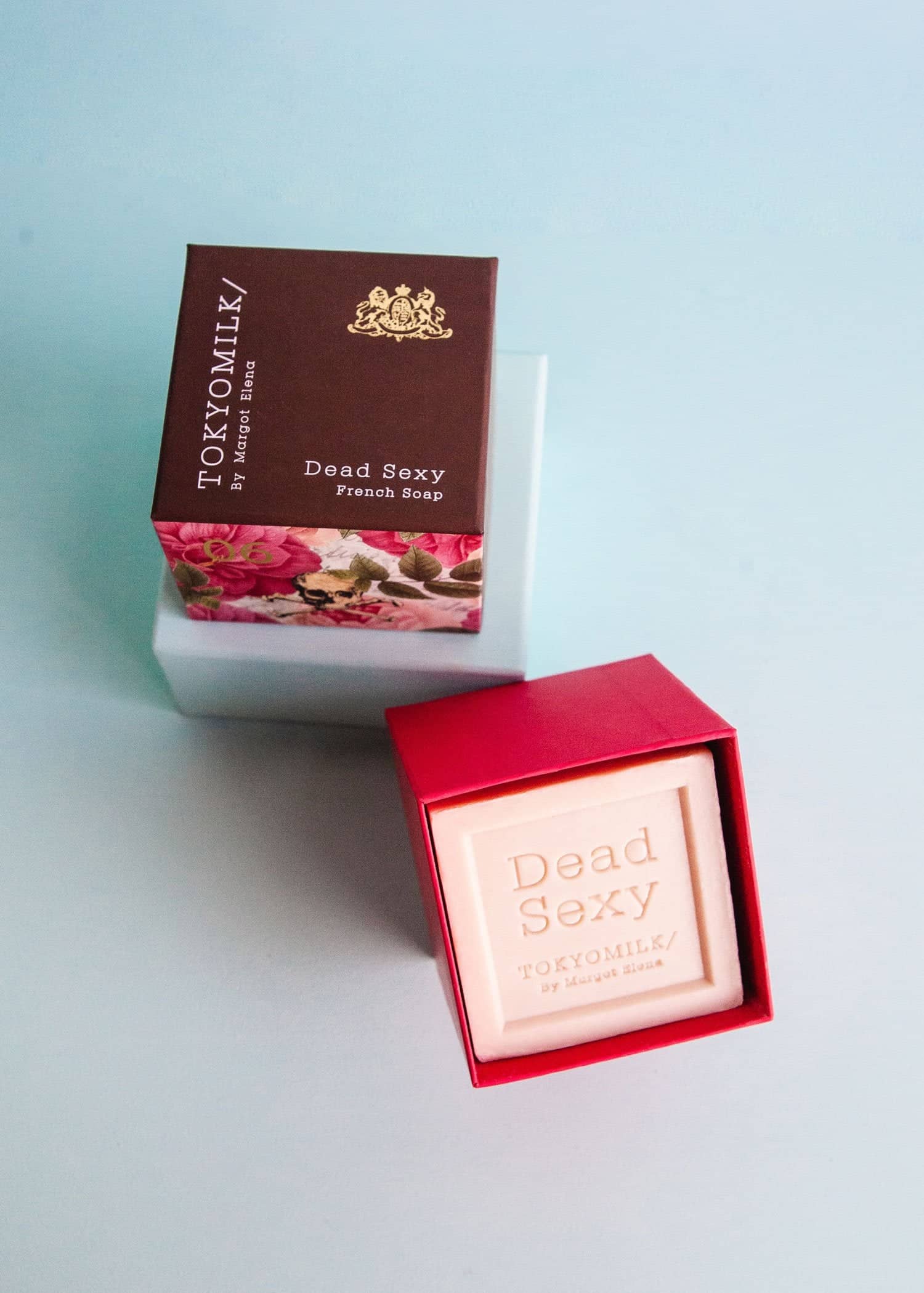 Product photograph of two TOKYOMILK by Margot Elena Dead Sexy French Soap boxes on a soft blue background, featuring a closed brown box with a floral skull pattern and an open bright red box revealing a pale pink embossed soap bar